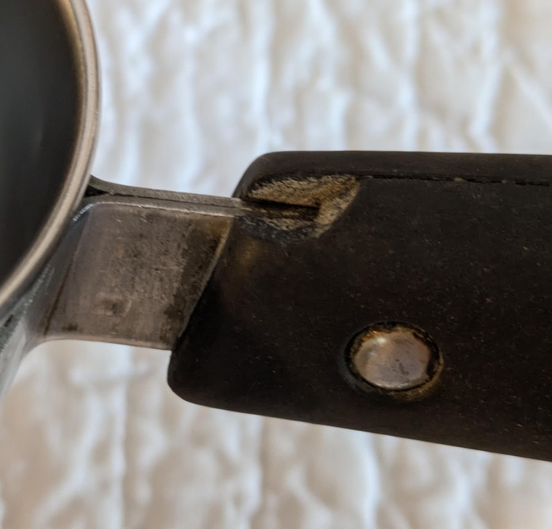 May include: Close-up of a metal and black handle attached to a stainless steel container. The handle is made of black material with a silver rivet. The metal shows signs of wear.
