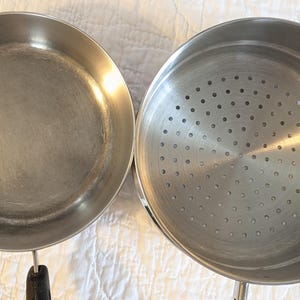 May include: Two stainless steel cooking pans. One is a frying pan with a black handle. The other is a steamer pot with a perforated base. Both pans are round and have a shiny, metallic finish.