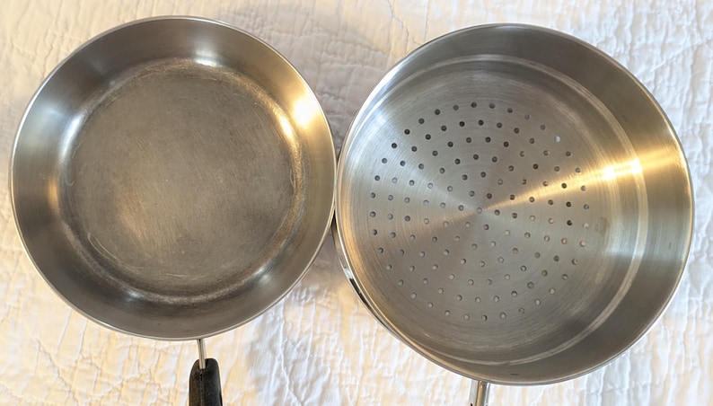 May include: Two stainless steel cooking pans. One is a frying pan with a black handle. The other is a steamer pot with a perforated base. Both pans are round and have a shiny, metallic finish.
