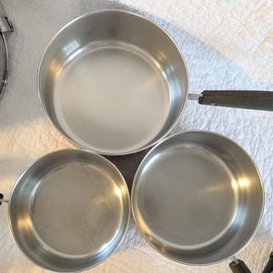 May include: A collection of three stainless steel saucepans. The pans are silver with black handles. The largest pan has a long handle, while the smaller pans have short handles. The pans are empty and on a white surface.