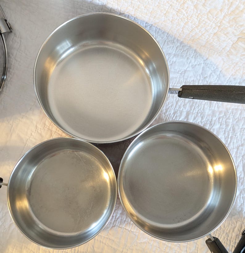 May include: A collection of three stainless steel saucepans. The pans are silver with black handles. The largest pan has a long handle, while the smaller pans have short handles. The pans are empty and on a white surface.