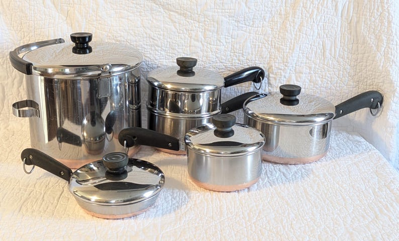 May include: A collection of stainless steel cooking pots and pans with black handles and lids. The set includes a large stockpot, a saucepan, and smaller pans. The bases of the pans have a copper-coloured trim.