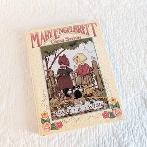 May include: A vintage "Mary Engelbreit Cross-Stitch" book. The cover features a cross-stitch design of two children looking over a fence, with the words "MAKE A WISH" below. The book has a cream-colored background with floral accents.