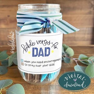 Op de afbeelding: Een heldere glazen pot met een blauw en wit lint om de bovenkant. De pot heeft een label met de tekst "Bible verses for DAD" met tekst eronder. Een digitale downloadsticker staat rechtsonder.