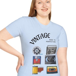Puede incluir: Camiseta azul claro con la palabra "VINTAGE" y la frase "AGED TO PERFECTION". La camiseta presenta ilustraciones de tecnología retro: una bobina de película, un disquete, un radiocasete, una cinta de casete y una cinta de 8 pistas. Un diseño nostálgico.