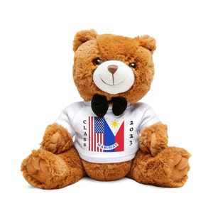 May include: A brown teddy bear wearing a white shirt with the American and Philippine flags and the text "CLASS 2023 MABUHAY". The bear is wearing a black bow tie.