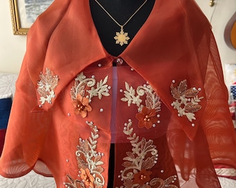 May include: A sheer burnt orange bolero jacket featuring a wide collar and intricate gold floral embroidery. The garment is adorned with small white pearls and three-dimensional fabric flowers, paired with a gold sunburst pendant necklace.