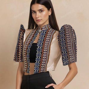 May include: A cream-colored, cropped top with patterned sleeves and a high collar. The sleeves and collar feature a geometric design in navy blue, brown, and white. The top is paired with black pants, creating a stylish ensemble.