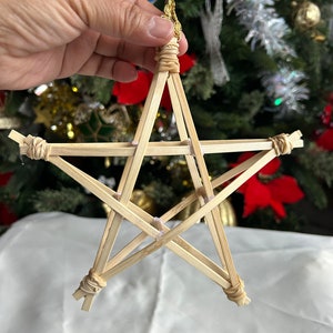 May include: A handcrafted Christmas star ornament made from thin bamboo strips, tied together with twine. The star is held by a gold-colored string, ready to hang on a Christmas tree. The background shows a decorated Christmas tree.