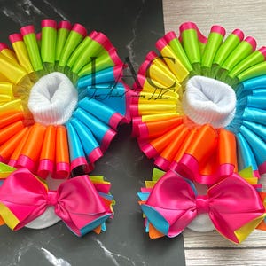 May include: A pair of white baby socks with colorful rainbow ruffles and pink bows. The ruffles are made of satin ribbon in shades of yellow, orange, red, pink, blue, green, and turquoise.