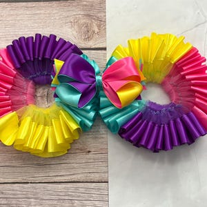 May include: Two circular hair accessories featuring ruffled layers of satin ribbon in vibrant shades of purple, hot pink, yellow, and turquoise. Each accessory is topped with a multi-colored bow made of purple, pink, yellow, and turquoise satin ribbon.