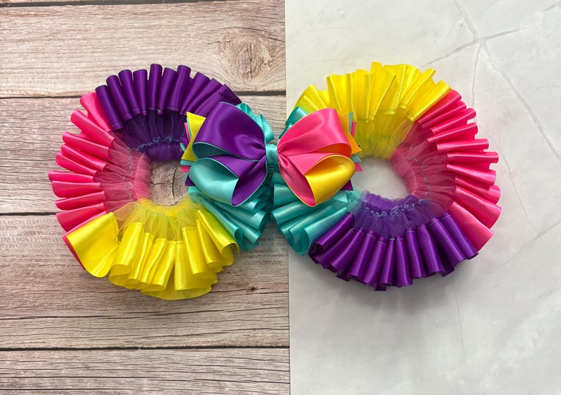 May include: Two circular hair accessories featuring ruffled layers of satin ribbon in vibrant shades of purple, hot pink, yellow, and turquoise. Each accessory is topped with a multi-colored bow made of purple, pink, yellow, and turquoise satin ribbon.