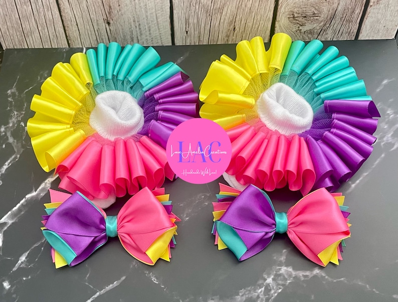 May include: Two pairs of baby socks with colorful, layered ribbon trim in a rainbow of colors: yellow, turquoise, purple, pink. Each pair has a matching bow with the same color scheme. The socks are on a dark surface.