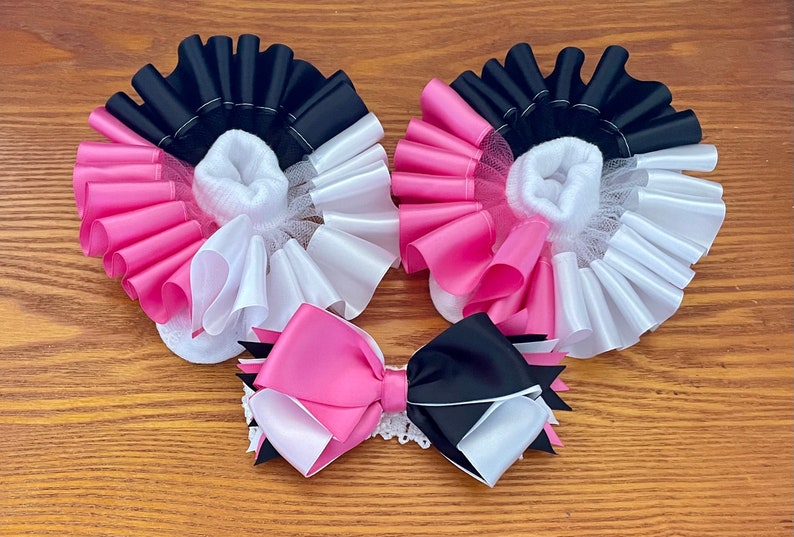 May include: A pair of white baby socks with pink, black, and white ruffled trim. A pink, black, and white bow is also included.
