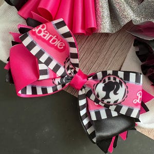 Peut inclure: Un n&oelig;ud rose et noir avec un ruban ray&eacute; blanc et noir sur lequel est inscrit le mot "Barbie" et une silhouette de poup&eacute;e Barbie.