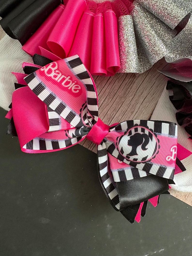 Peut inclure: Un n&oelig;ud rose et noir avec un ruban ray&eacute; blanc et noir sur lequel est inscrit le mot "Barbie" et une silhouette de poup&eacute;e Barbie.