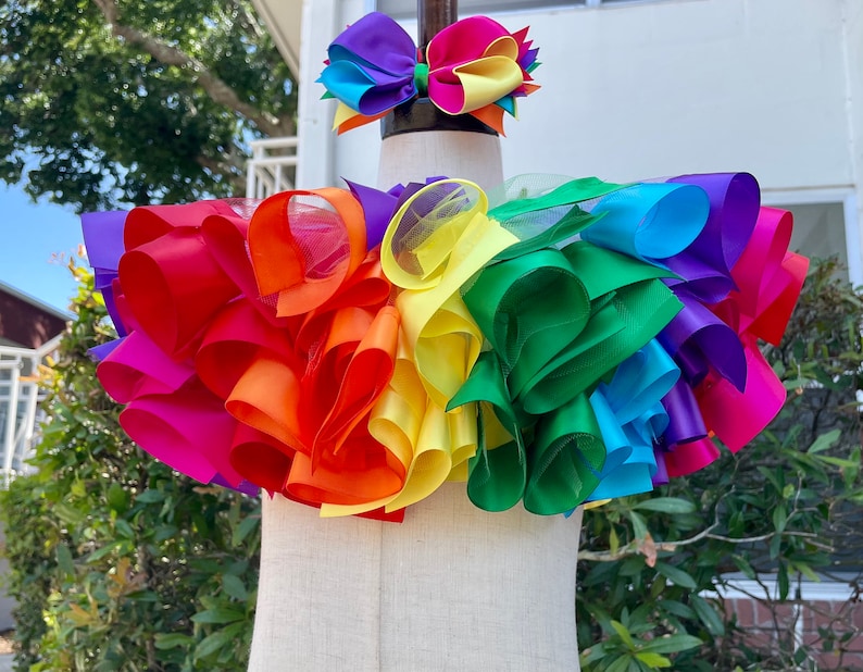 May include: Rainbow layered tulle and ribbon skirt with a matching rainbow ribbon hair bow. The skirt is made of multiple layers of tulle and ribbon in the colors of the rainbow. The hair bow is made of a single layer of ribbon in the colors of the rainbow.