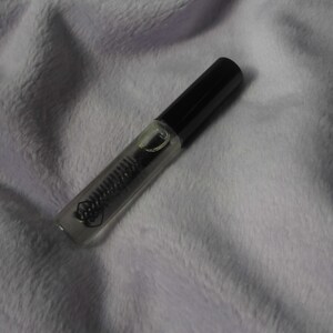 May include: A clear glass tube with a black cap containing a clear liquid. The tube is lying on a white fuzzy surface.