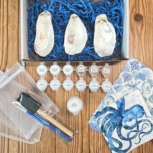 May include: An open cardboard box with three oyster shells, blue paper filler, paint pots, brushes, and napkins. The napkins feature an octopus and seashell designs. The paint colors include white, gold, and satin finishes.