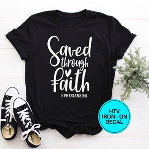 May include: Black t-shirt with white text that reads "Saved through faith Ephesians 2:8". The shirt is laying flat on a white surface with a pair of black and white sneakers and a green plant in the background. The image also includes a blue circle with white text that reads "HTV IRON-ON DECAL".