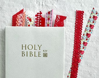Puede incluir: Una Biblia blanca con letras doradas y un pequeño símbolo dorado. La Biblia tiene varios marcadores de cinta roja, rosa y blanca.