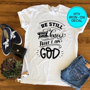 May include: White t-shirt with black text that reads "Be Still and Know That I Am God". The shirt is laid flat on a wooden surface with a pair of blue sneakers, a small potted plant, and a pair of sunglasses.