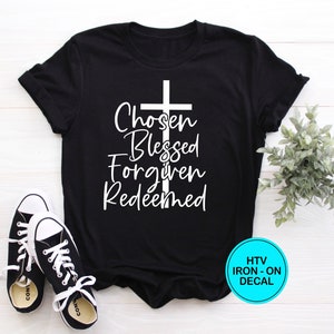 May include: Black t-shirt with a white cross and the words "Chosen Blessed Forgiven Redeemed". The shirt is laying flat on a white wooden surface with a small green plant and two black sneakers.
