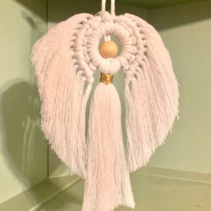 以下が含まれることがあります： 木製のビーズの頭と金色のトリムが付いた白いマクラメの天使のオーナメント。天使は長く流れるような羽根とタッセルスカートを持っています。