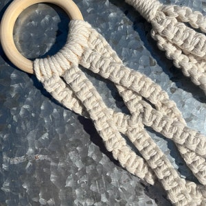 May include: A macrame plant hanger with a wooden ring. The hanger is made of natural cotton rope and has a simple, minimalist design.