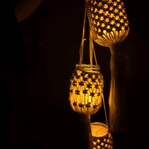 May include: Three hanging mason jar lanterns with a woven macrame design. The lanterns are lit from within, casting a warm yellow glow. Each lantern has a tassel at the bottom and is suspended by a string.