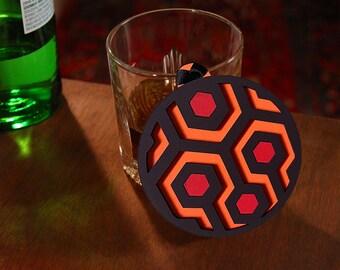May include: A black, orange, and red coaster with a geometric pattern, resembling a honeycomb or a stylized design. The coaster sits next to a glass of amber liquid and a green bottle. The coaster has a black ribbon attached.