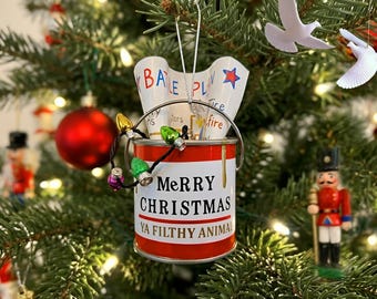 May include: A red and white paint can Christmas ornament featuring the text Merry Christmas Ya Filthy Animal. Inside the can is a paper scroll labeled My Battle Plan with a blue star. A string of miniature multicolored holiday lights is attached.