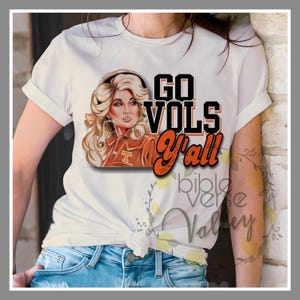 Puede incluir: Camiseta blanca con un gráfico de una mujer con cabello rubio que lleva una chaqueta roja y naranja con el texto "GO VOLS Y'all" en letras negras y naranjas.