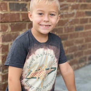 Puede incluir: Un ni&ntilde;o peque&ntilde;o con una camiseta gris oscuro con un dise&ntilde;o blanqueado que presenta el texto "Good ole Rocky Top" y un gr&aacute;fico del estado de Tennessee. El dise&ntilde;o es en naranja, amarillo y negro.