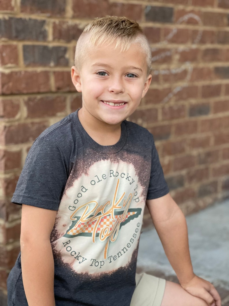 Puede incluir: Un ni&ntilde;o peque&ntilde;o con una camiseta gris oscuro con un dise&ntilde;o blanqueado que presenta el texto "Good ole Rocky Top" y un gr&aacute;fico del estado de Tennessee. El dise&ntilde;o es en naranja, amarillo y negro.
