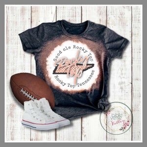Puede incluir: Una camiseta negra con un dise&ntilde;o blanqueado que presenta el texto "Good ole Rocky Top" y un contorno a cuadros del estado de Tennessee. La camiseta se combina con un bal&oacute;n de f&uacute;tbol marr&oacute;n y zapatillas blancas de ca&ntilde;a alta.