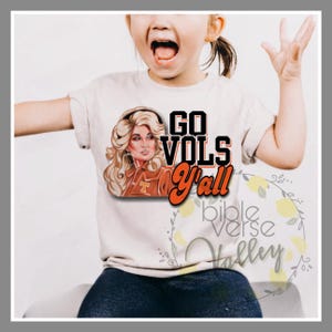 Puede incluir: Una camiseta blanca con un gráfico de una mujer con cabello rubio y una chaqueta roja y naranja. El texto "GO VOLS Y'all" está impreso en naranja en la camiseta.