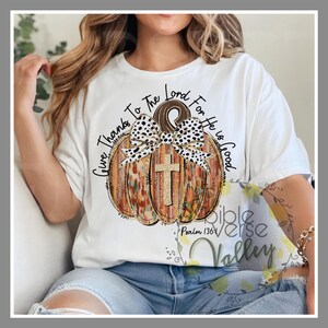 Puede incluir: Camiseta blanca con un gráfico de una calabaza marrón y naranja con un lazo de lunares blanco y negro y una cruz dorada. El texto "Give Thanks To The Lord For He Is Good" está escrito alrededor de la calabaza. El texto "Psalm 136:1" está escrito debajo de la calabaza.