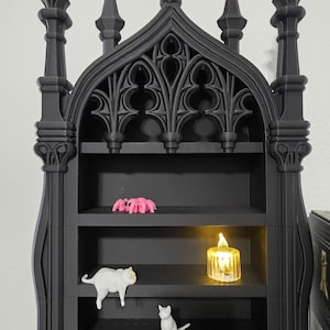 May include: A black gothic-style shelf unit with three shelves. The top of the unit has decorative spires and arches. The shelves hold a pink spider figurine, a white cat figurine, a small white cat figurine, and a lit candle.