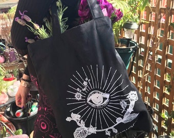 May include: Black tote bag with a white design of an eye with a sunburst and a floral motif. The text "Mystic Vision" is visible at the bottom of the design.