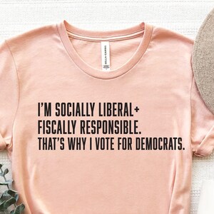 May include: A peach-colored t-shirt with the text "I'M SOCIALLY LIBERAL+ FISCALLY RESPONSIBLE. THAT'S WHY I VOTE FOR DEMOCRATS."