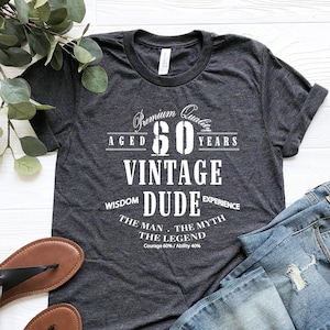 May include: A dark gray t-shirt with white text that reads "Premium Quality Aged 60 Years Vintage Dude Wisdom Experience The Man. The Myth. The Legend. Courage 60% / Ability 40%"