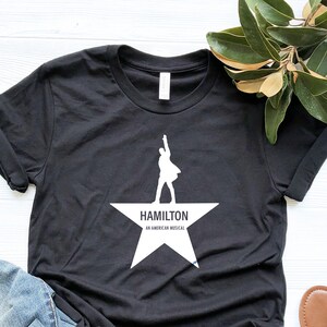 May include: Black t-shirt with a white silhouette of a person standing on a white star with the text "HAMILTON AN AMERICAN MUSICAL".