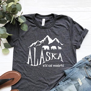 以下が含まれることがあります： 暗いヘザーグレーのTシャツに、白い山と熊のグラフィックがプリントされています。グラフィックの下には「ALASKA wild and wonderful」というテキストがプリントされています。