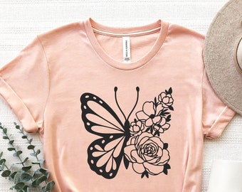 May include: A light pink t-shirt with a black butterfly and floral design. The butterfly is in flight and has its wings spread out. The flowers are arranged around the butterfly and are in bloom.