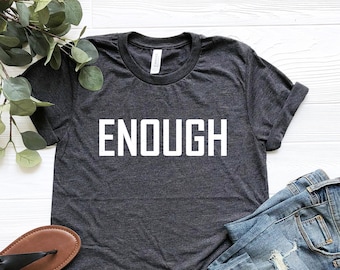Może przedstawiać: Szary t-shirt z nadrukiem "ENOUGH" w kolorze białym na przodzie. Koszulka leży płasko na białym tle z parą brązowych sandałów i niebieskimi dżinsami.