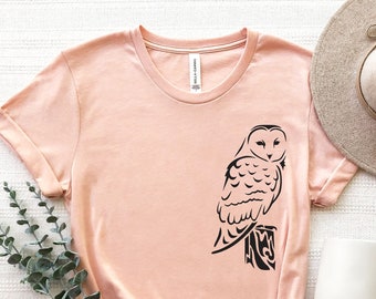 May include: A peach-colored t-shirt with a black silhouette of an owl perched on a branch. The owl has a detailed design with feathers and a round head.