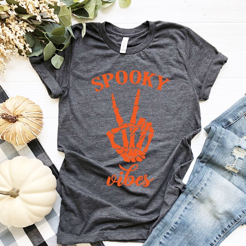 May include: Dark gray t-shirt with the words "SPOOKY vibes" and a skeleton hand making a peace sign in orange. The shirt is paired with blue jeans and decorative pumpkins.