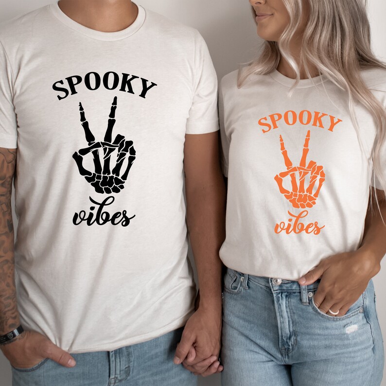 May include: Two cream-colored t-shirts with the words "SPOOKY vibes" printed on them. One shirt has black text and a black skeleton hand making a peace sign. The other shirt has orange text and an orange skeleton hand.