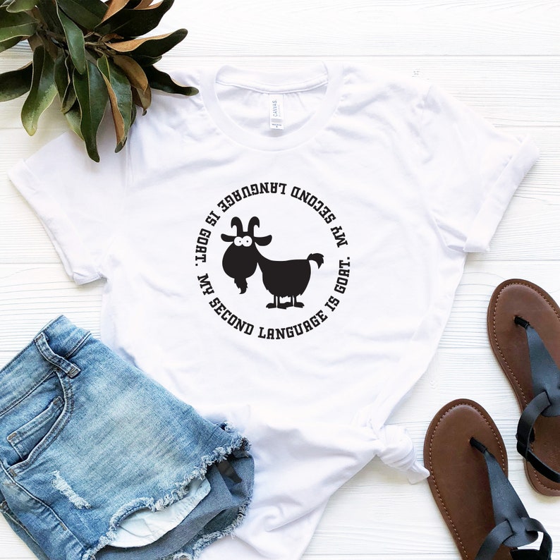 May include: White t-shirt with a black circle graphic featuring a cartoon goat and the text "My second language is goat.  My second language is goat."